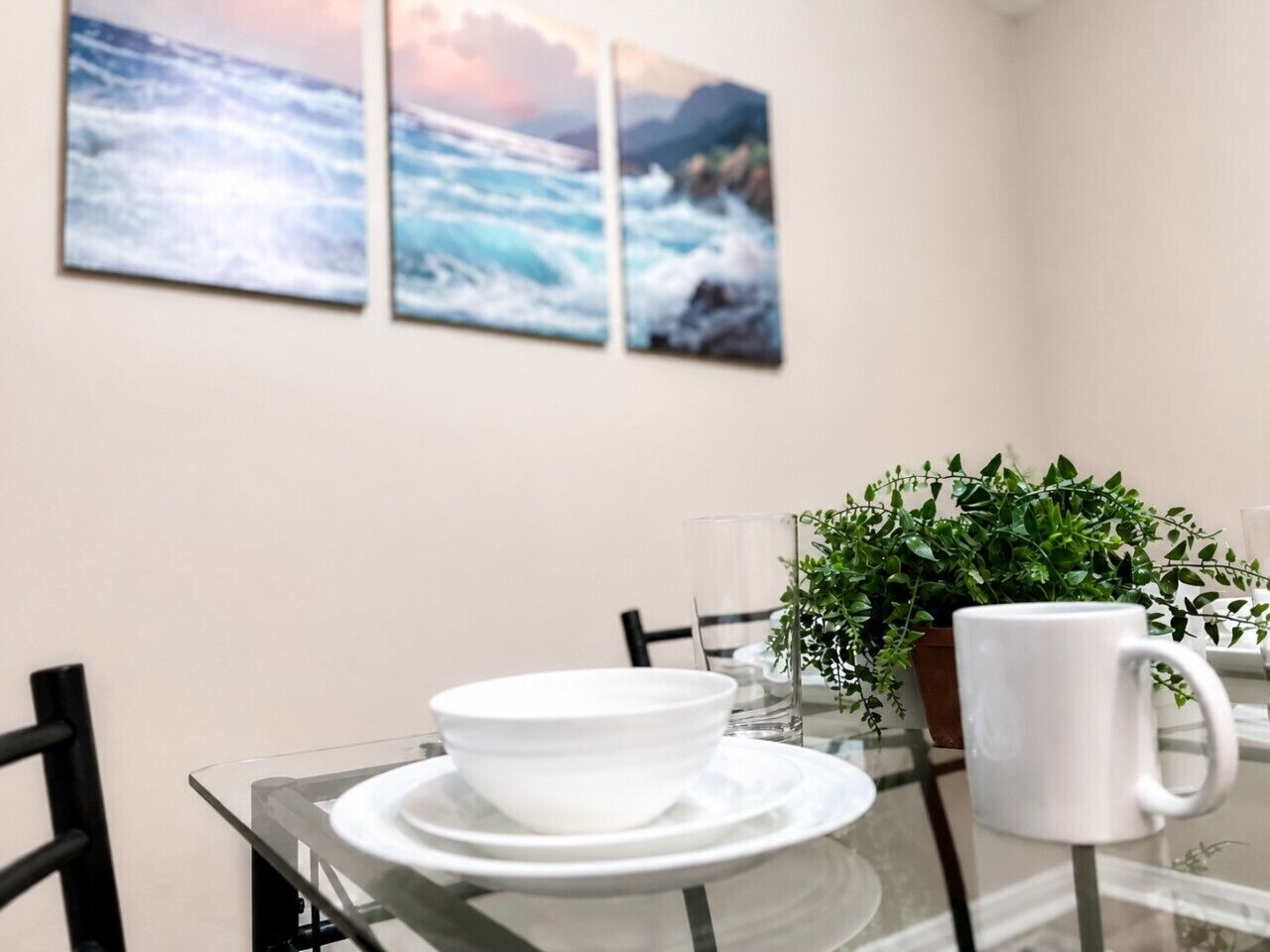 A dining area with a glass table, white plates, bowls, and a white mug. There are chairs around the table and a potted plant in the background.