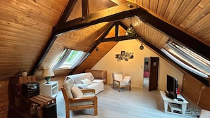 A cozy attic room with a wooden ceiling, a sofa, a chair, a television, and a small table. - Charming Breton House Near Petit Havre Beach (Pordic)
