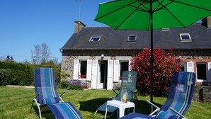 A stone house with a sloped roof, two lounge chairs, a green umbrella, and a small table with a bottle. - Charming Breton House Near Petit Havre Beach (Pordic)