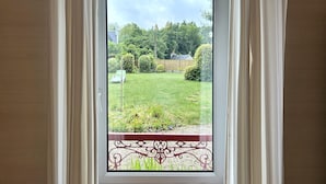 A view through a window with white curtains, revealing a backyard with a lawn, a green chair, and a decorative metal fence. - Charming Breton House Near Petit Havre Beach (Pordic)