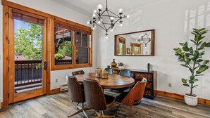 A dining area with a round table, chairs, a chandelier, a potted plant, and a large window with a view of trees.