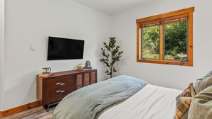 A bedroom with a bed, a dresser, a flat-screen TV, a window with a view of greenery, and a ceiling light.