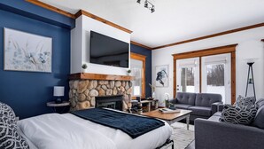 A modern bedroom with a stone fireplace, a large bed, a TV mounted above the fireplace, and a seating area with a sofa and armchairs. - Snowbridge (Blue Mountains)