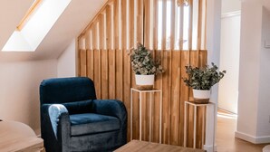 Ein modernes Wohnzimmer mit schrägverdeckter Decke, einem blauen Sessel, zwei Topfpflanzen und einem Holztrennfeld.