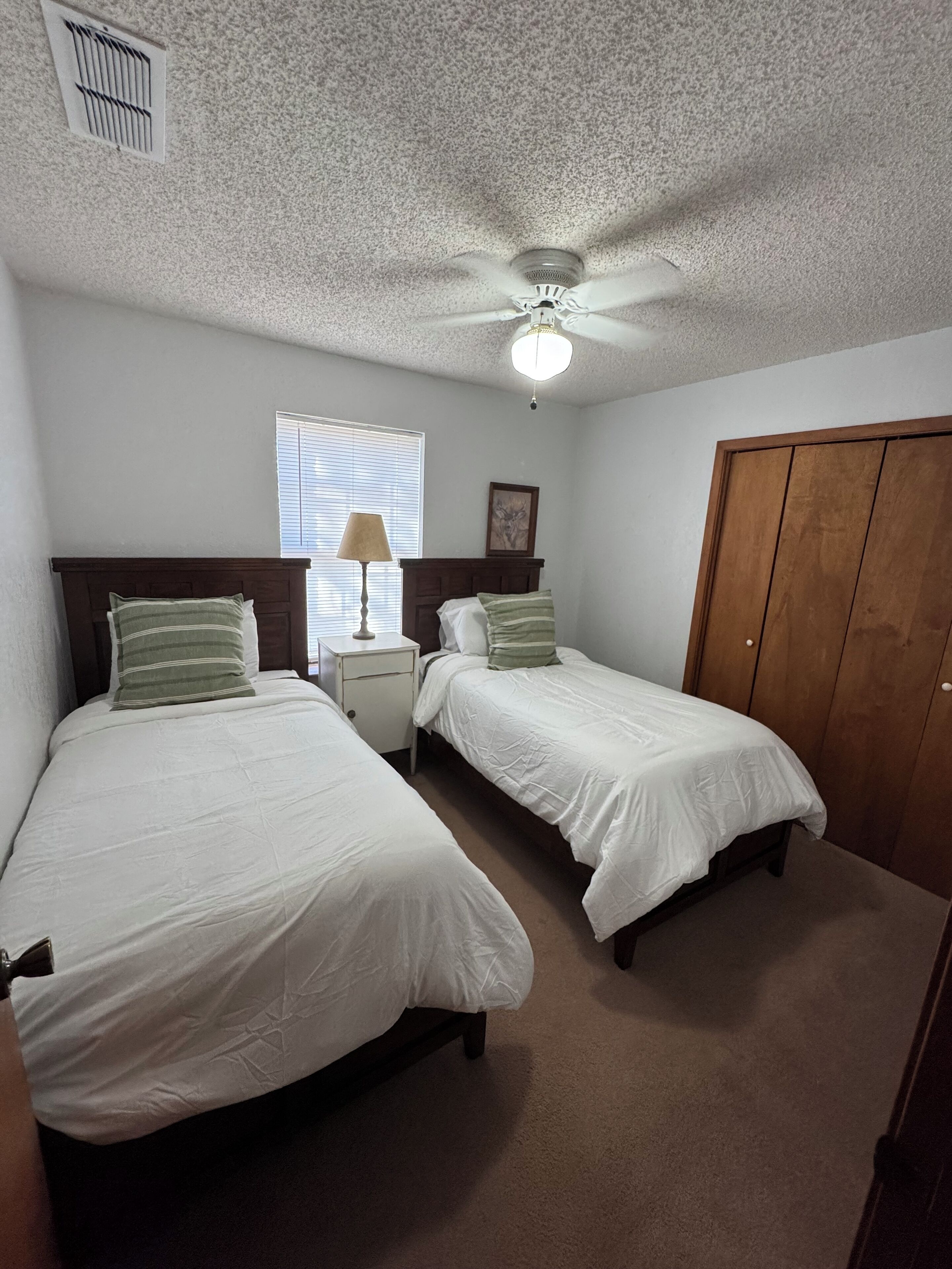 A bedroom with two single beds, a nightstand, a lamp, a ceiling fan, a window with blinds, and a wooden wardrobe.