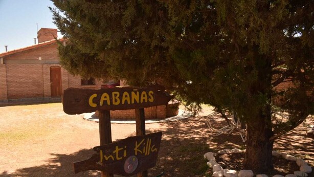 Een houten bord met de tekst 'CABANAS' en 'Loti Killa', buiten geplaatst, met een boom en een bakstenen gebouw op de achtergrond.