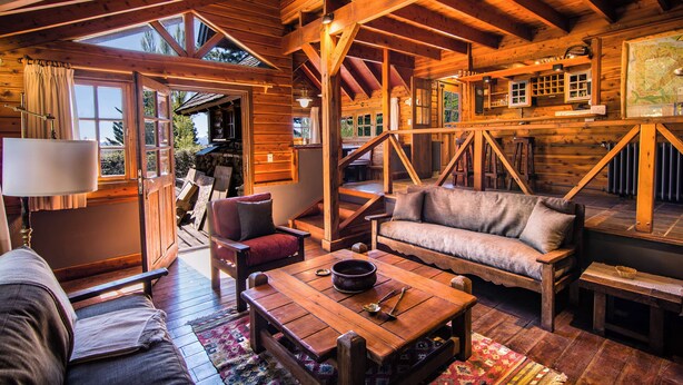 A cozy log cabin interior with a wooden staircase, a sofa, a coffee table, and a dining table.
