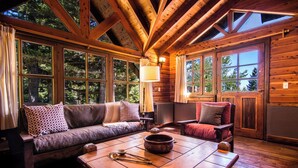 A cozy cabin interior with a wooden floor, a sofa, a table with a bowl and utensils, and large windows offering a view of trees.