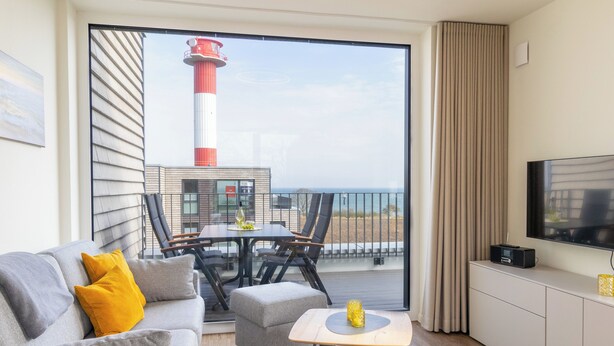 A modern living room with a grey sofa, a small ottoman, a coffee table, and a flat-screen TV on a white cabinet. A dining area with a table and chairs is visible through a large glass door, overlooking a beach and a lighthouse.