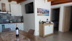 A modern kitchen with white cabinets, a TV mounted on the wall, and a wooden dining table with a bottle of wine.