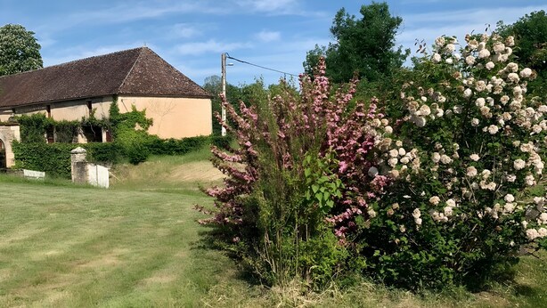 Una casa rural con techo inclinado, rodeada de un césped cuidado y arbustos florecientes.