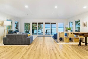A spacious living room with a grey sofa, wooden flooring, and large windows offering a view of the water.