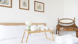 Een net opgemaakt bed met witte lakens, een houten nachtkastje met een dienblad waarop witte theekoppen en een bloevas staan, en een houten stoel met een spiegel.