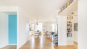 Een ruime, goed verlichte kamer met een houten vloer, een eethoek met stoelen en een tafel, en een boekenplank vol boeken.