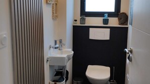 Un baño pequeño con un lavamanos blanco, un inodoro y una ventana sobre el inodoro. Hay un estante con un dispensador de jabón y una planta en maceta.