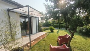 Una casa moderna con puerta de cristal, un porche cubierto y un césped cuidado con un árbol y dos sillas de plástico rojas.