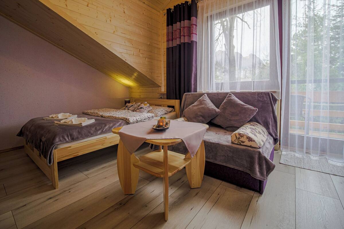 Une cabane chaleureuse avec un lit en mezzanine, un canapé, une petite table et une fenêtre avec des rideaux.