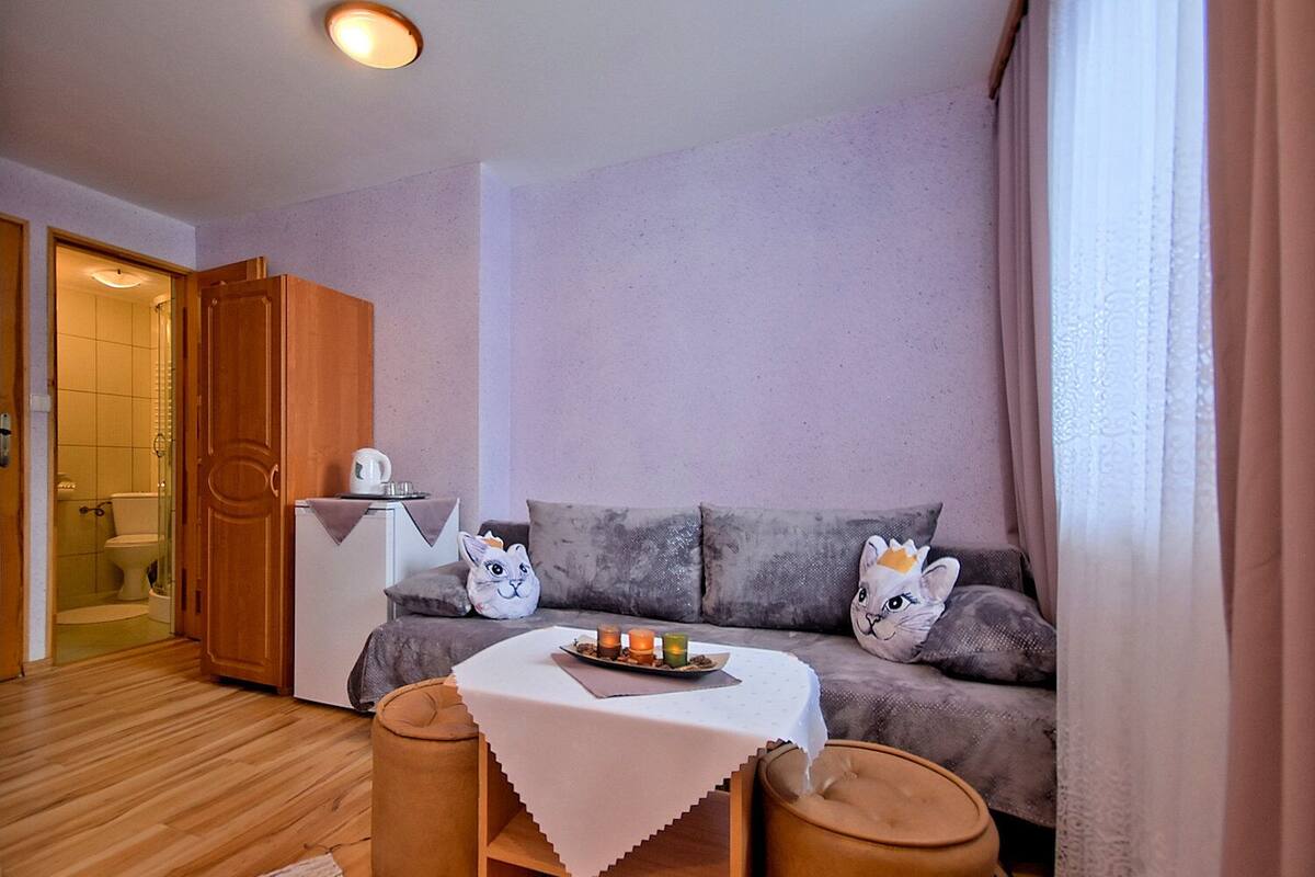 Un salon confortable avec un canapé, deux poufs, une petite table recouverte d’une nappe blanche et une porte donnant sur une salle de bain.