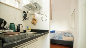 A compact kitchen with a sink, kettle, and coffee maker. A bed with a floral bedspread is visible in the adjacent room. - SUH Catete 02 Christ View (Rio de Janeiro)