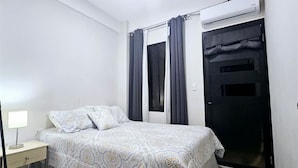 Un dormitorio con cama, mesita de noche, ventana con cortinas y equipo de aire acondicionado.