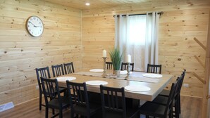 Una habitación con paneles de madera, una mesa redonda dispuesta para seis personas, sillas alrededor, un reloj en la pared y una ventana con cortinas.