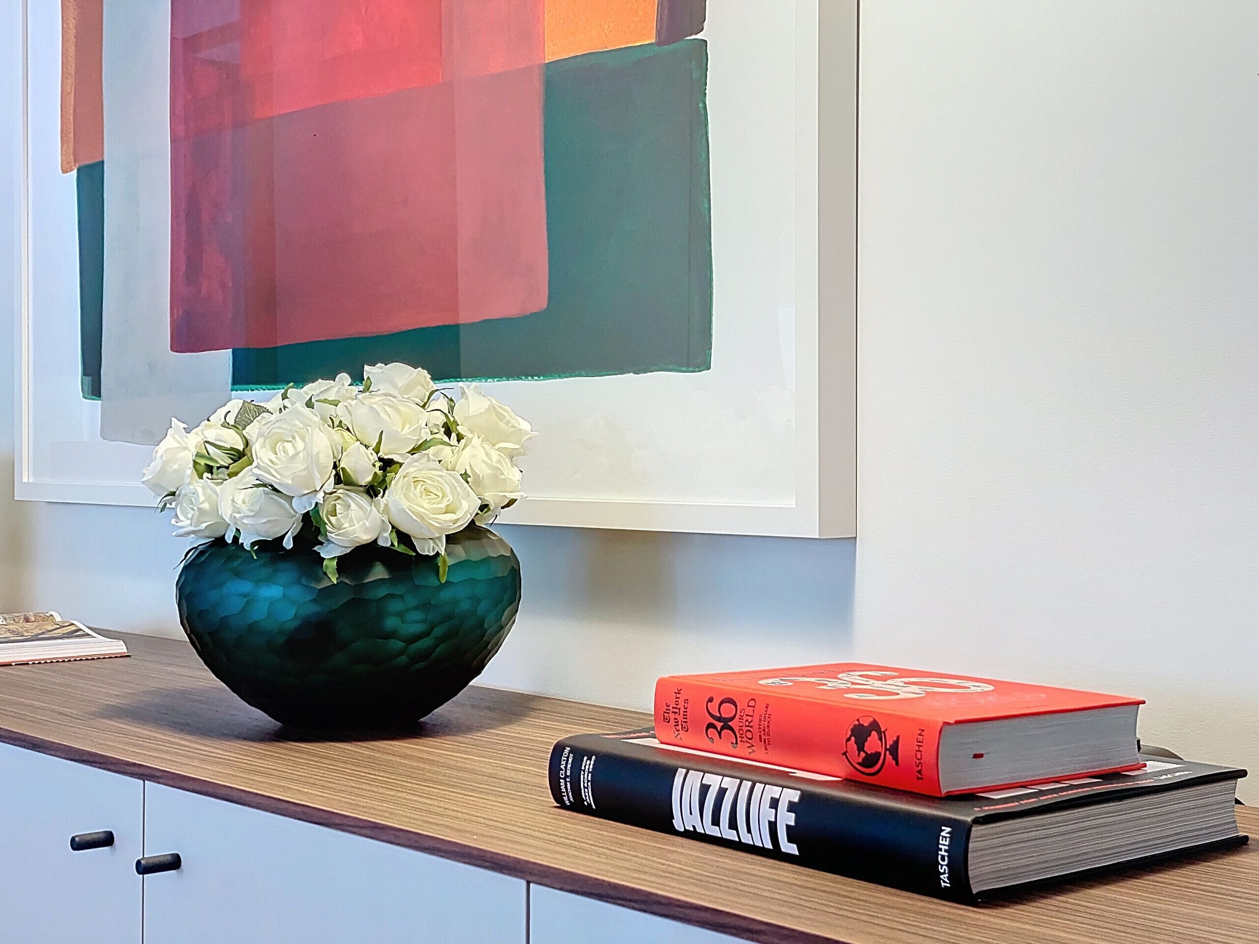 Un bureau sur lequel se trouve un vase vert contenant des fleurs blanches, un livre rouge intitulé « The New York Times » et un livre noir intitulé « JAZZ LIFE ».