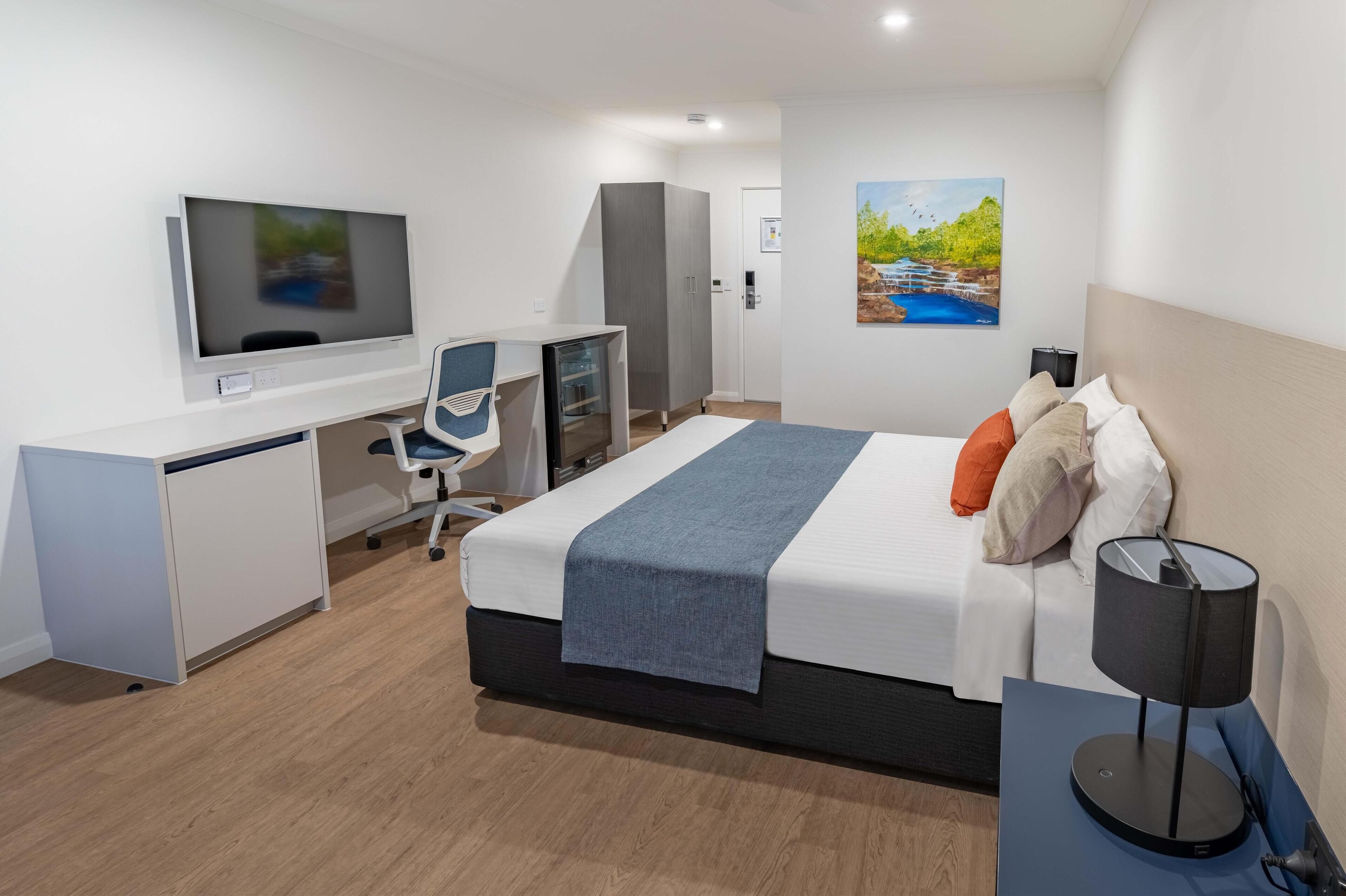 A modern hotel room with a bed, desk, chair, TV, wardrobe, and artwork on the wall.