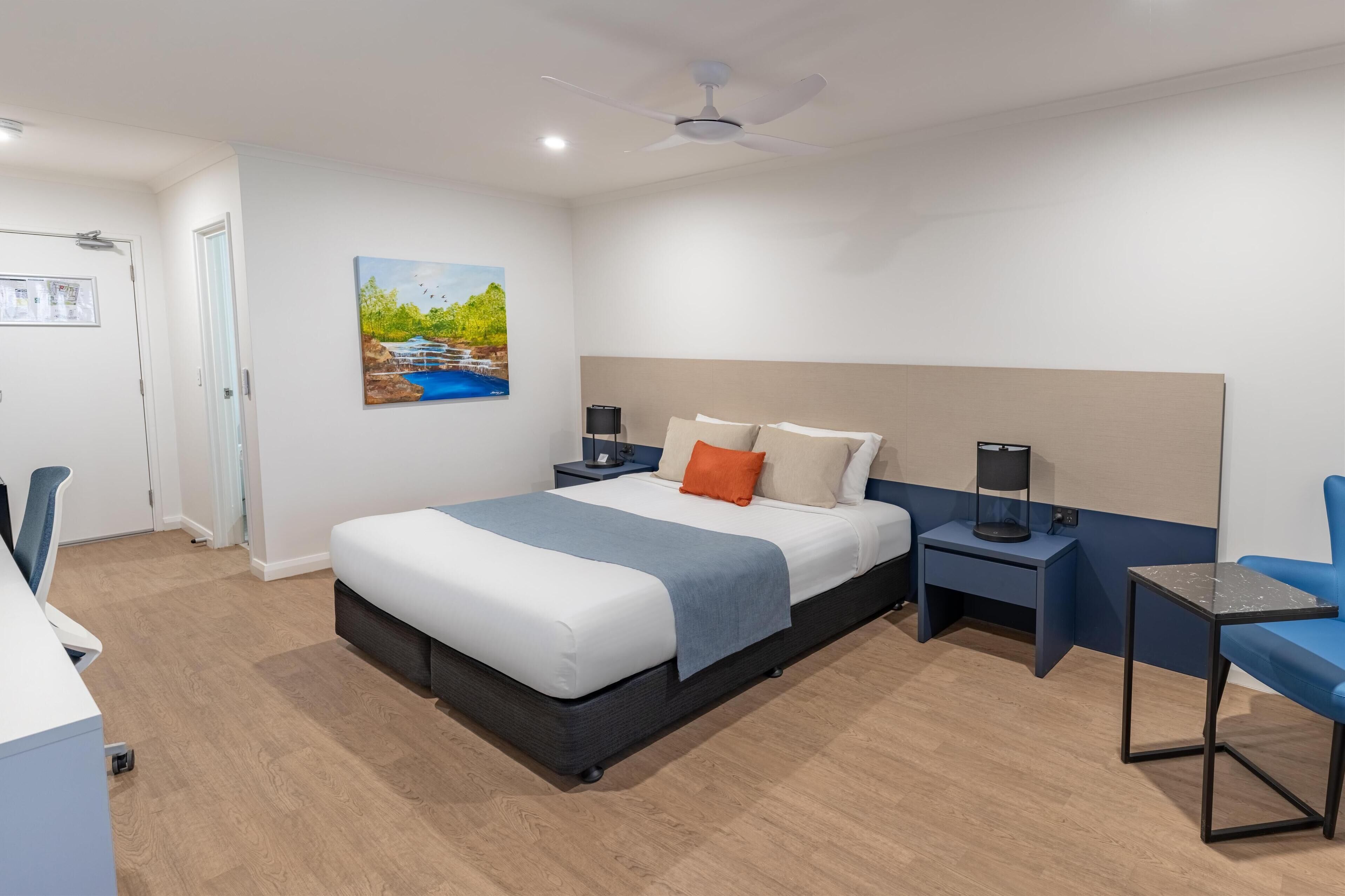 Une chambre d’hôtel moderne, équipée d’un lit, d’un bureau, d’une chaise, d’une table de chevet, d’une lampe et d’un tableau au mur.
