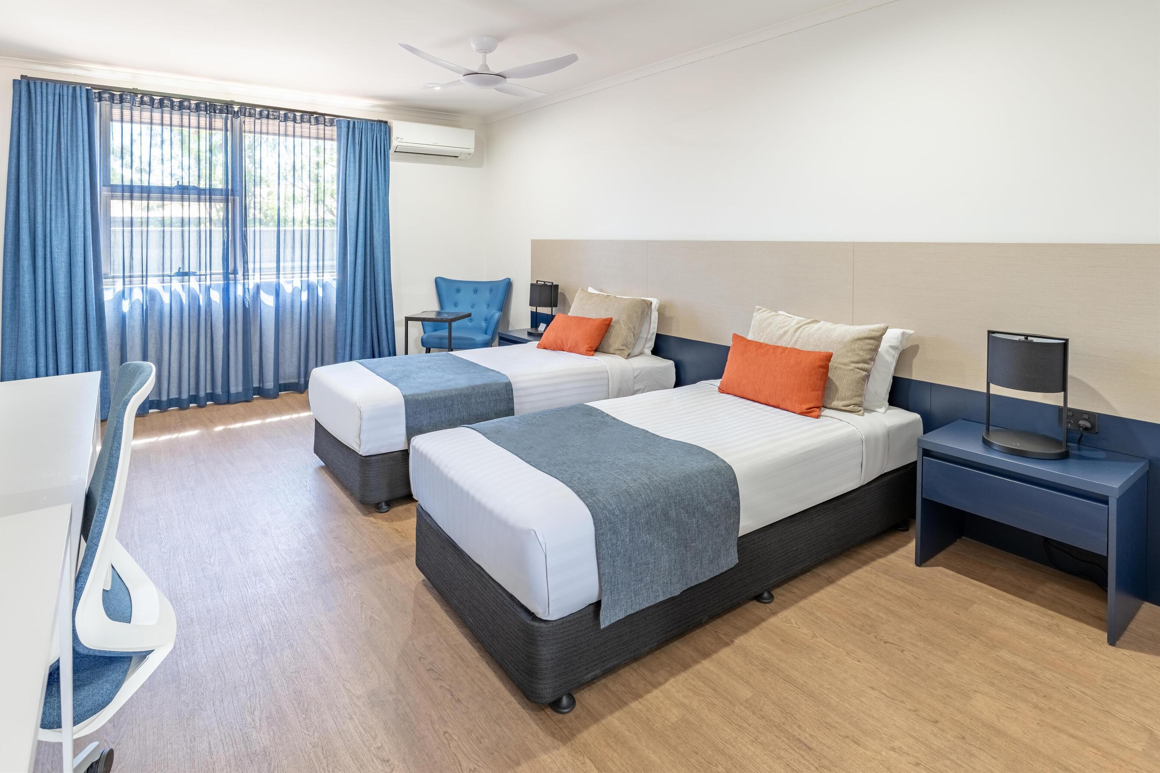 A hotel room with two beds, a desk, a chair, and a ceiling fan.