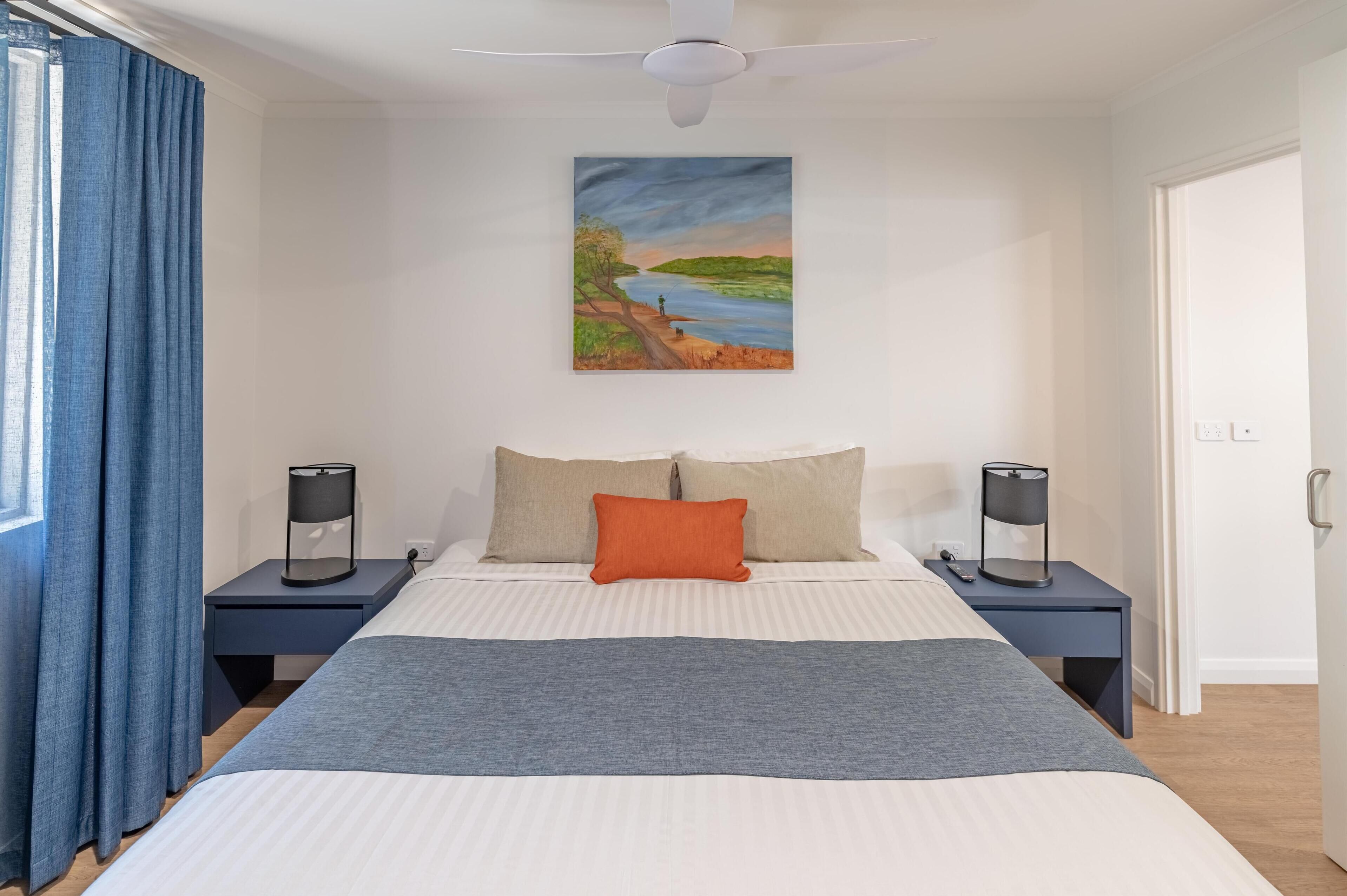 A bedroom with a bed, two nightstands, a ceiling fan, a painting, and a door.
