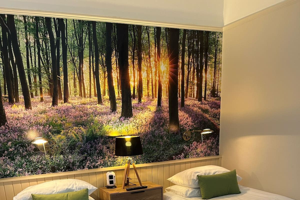 Une chambre avec deux lits, une table de chevet avec une lampe et une fresque murale représentant une scène de forêt.
