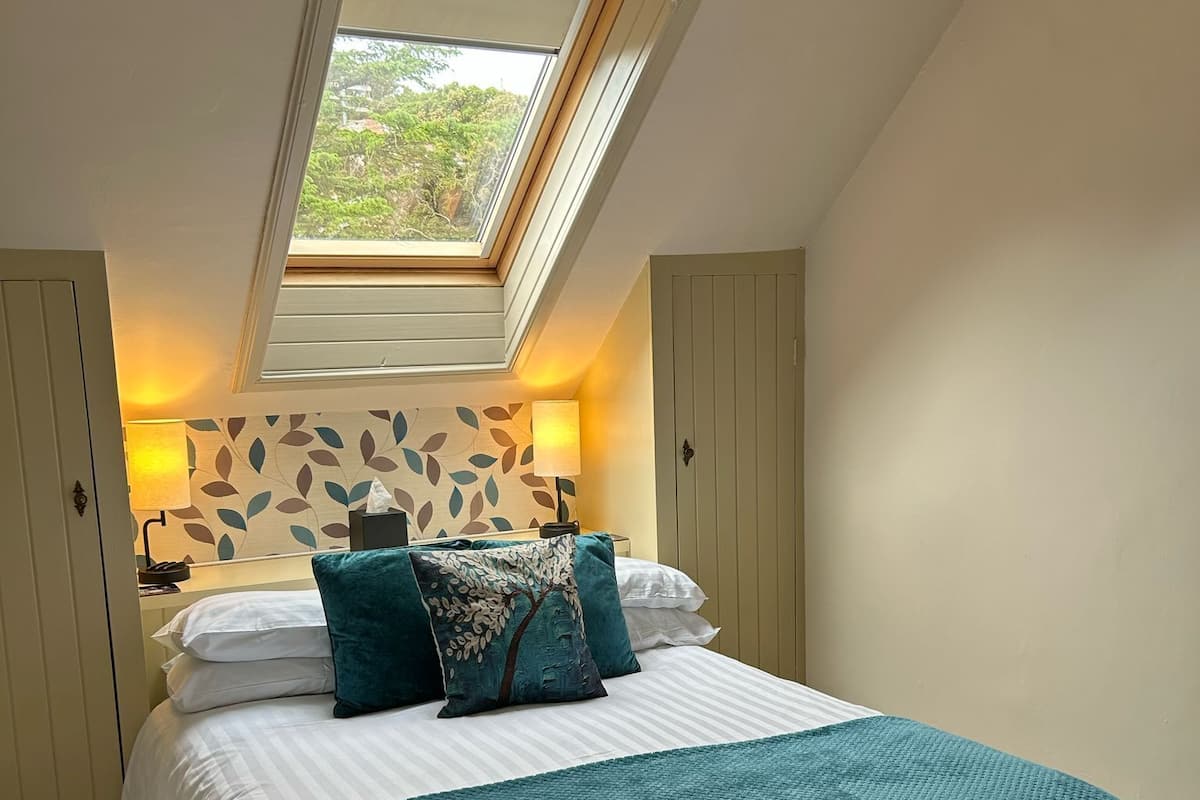Une chambre avec un puits de lumière, un lit avec une literie de couleur turquoise et un mur recouvert d’un papier peint à motifs de feuilles.