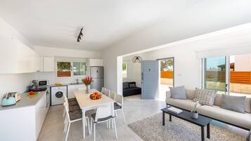 Una cocina moderna con armarios blancos, una zona de comedor con mesa y sillas, y una sala de estar con sofá y mesa de centro.