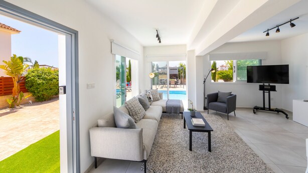 Una sala de estar moderna con un sofá, una mesa de centro y un televisor. Se ve una piscina a través de las puertas de cristal.