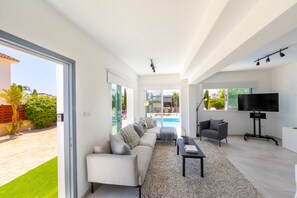 A modern living room with a sofa, a coffee table, and a TV. There is a pool visible through the glass doors. - Ayia Napa Marina Vibes (Sotira)