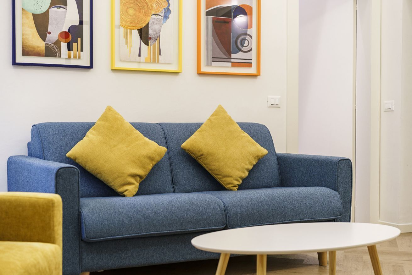 A blue sofa with two mustard-colored pillows, a white round table, and three framed artworks on the wall.