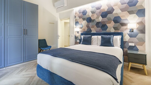 A modern hotel room with a large bed, a wardrobe, a chair, and a geometric-patterned headboard. - Designer Apartment in Oltrarno (Firenze)