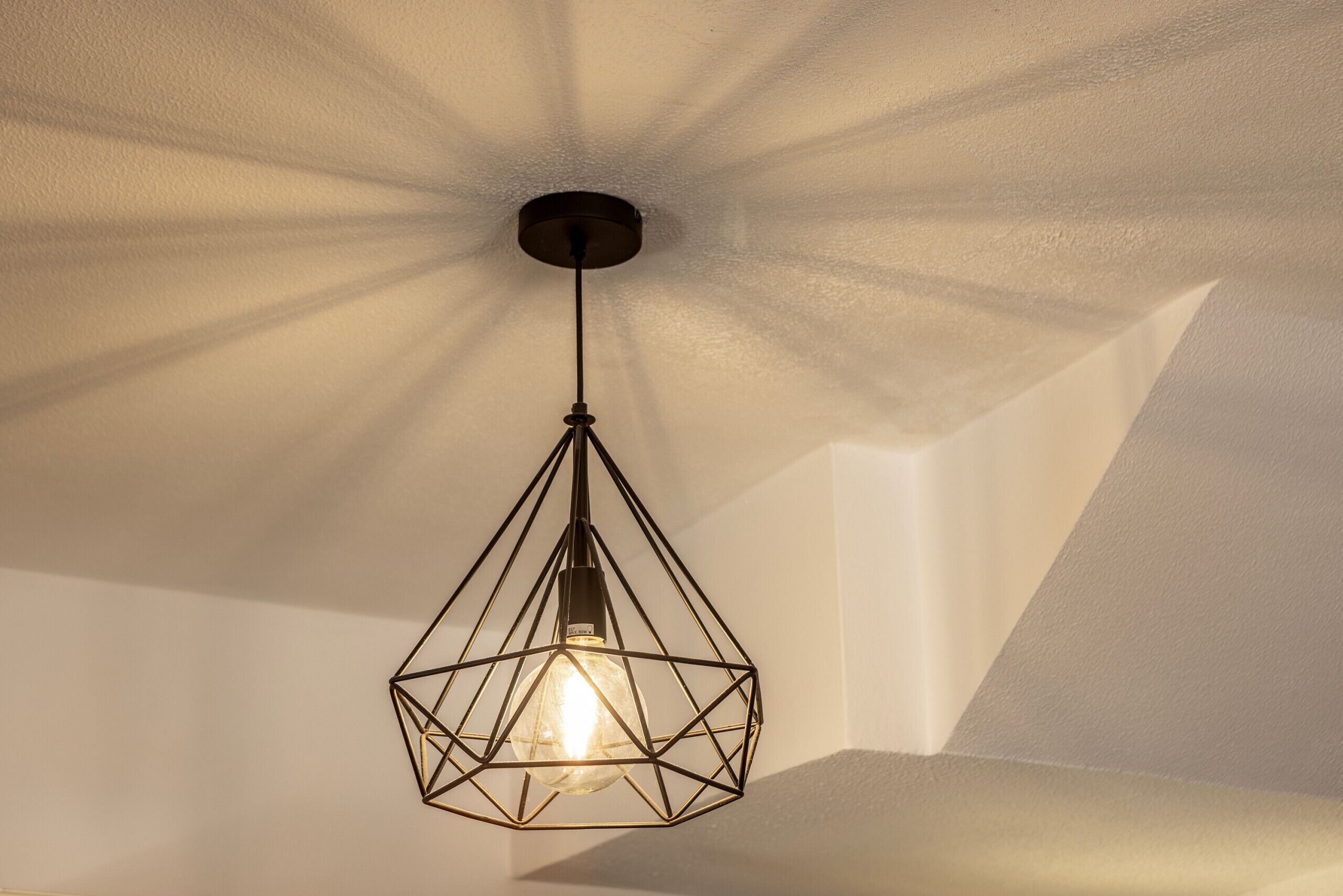 Een geometrische hanglamp met zichtbare gloeilamp, hangend aan een plafond met een structuur.