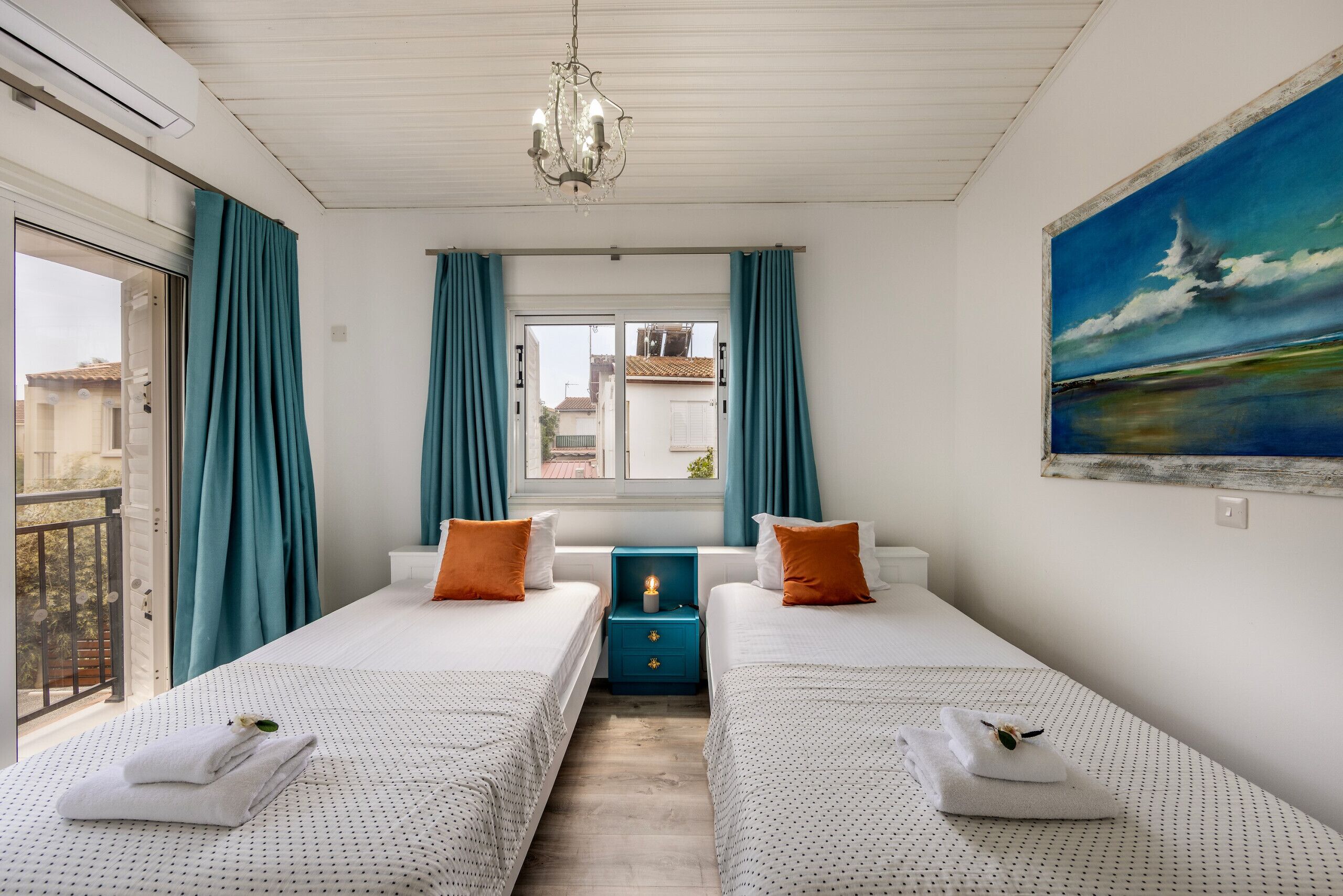 Habitación de hotel con dos camas, cortinas color turquesa, una araña, un cuadro con una vista marina y una ventana que ofrece vistas de edificios.