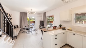 Una cocina moderna con armarios blancos, encimera de madera y horno empotrado. Hay un comedor con una mesa y sillas, y una escalera que sube a otro nivel.