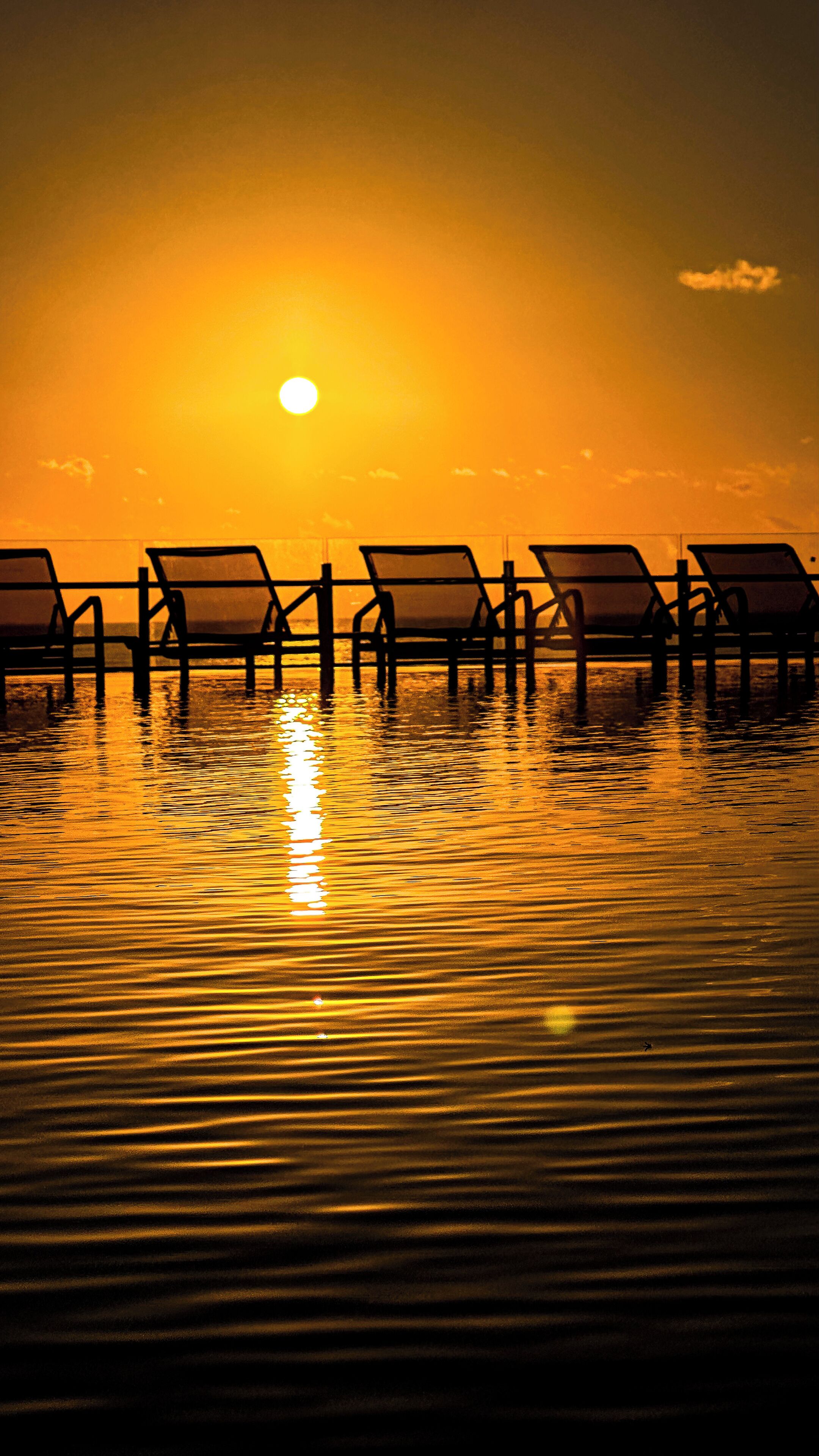 A sunset over a body of water with rows of lounge chairs aligned parallel to the water's edge.