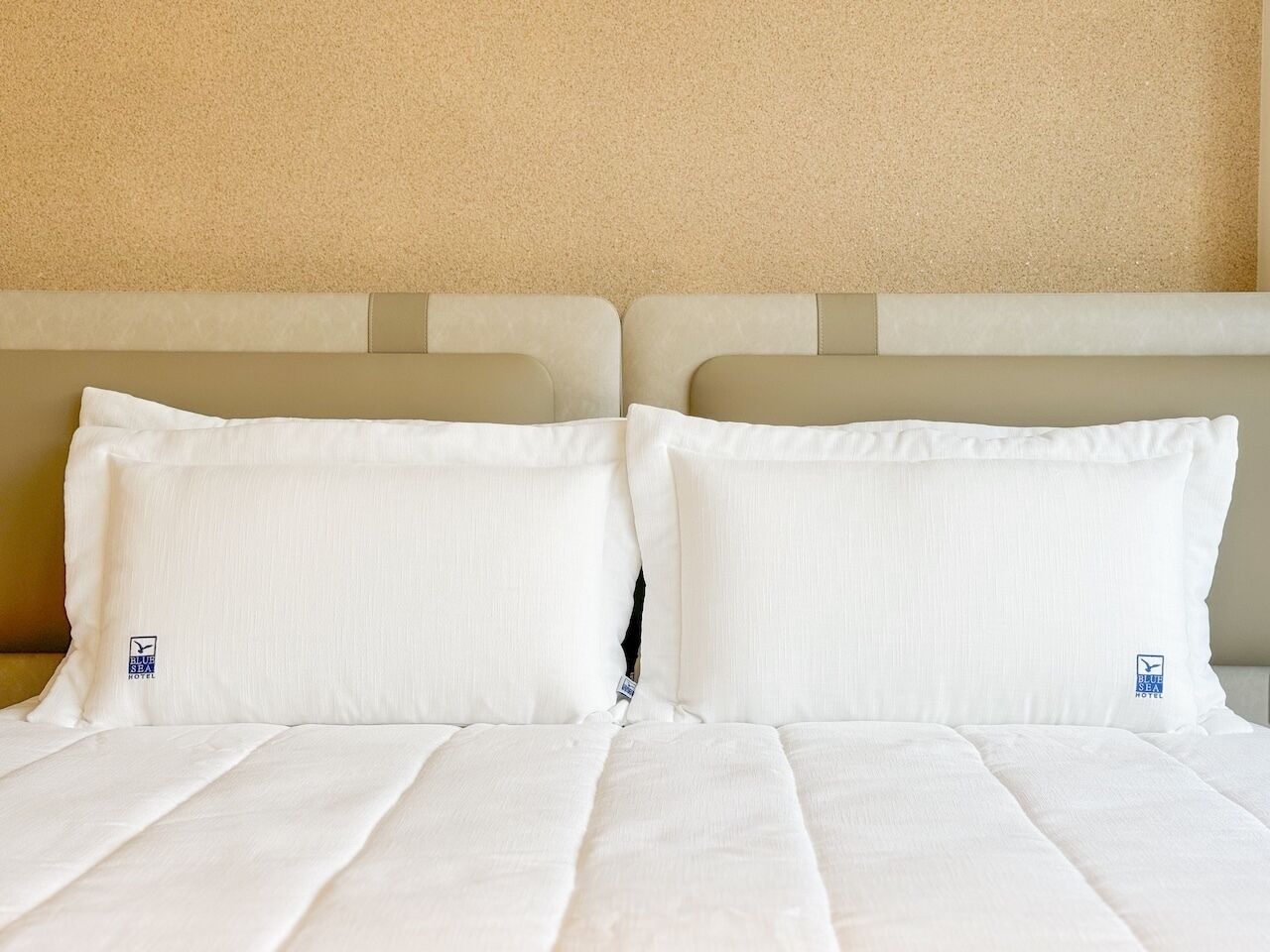 Una cama bien hecha con sábanas y almohadas blancas, un cabecero color beige y una pared texturizada en tono beige.