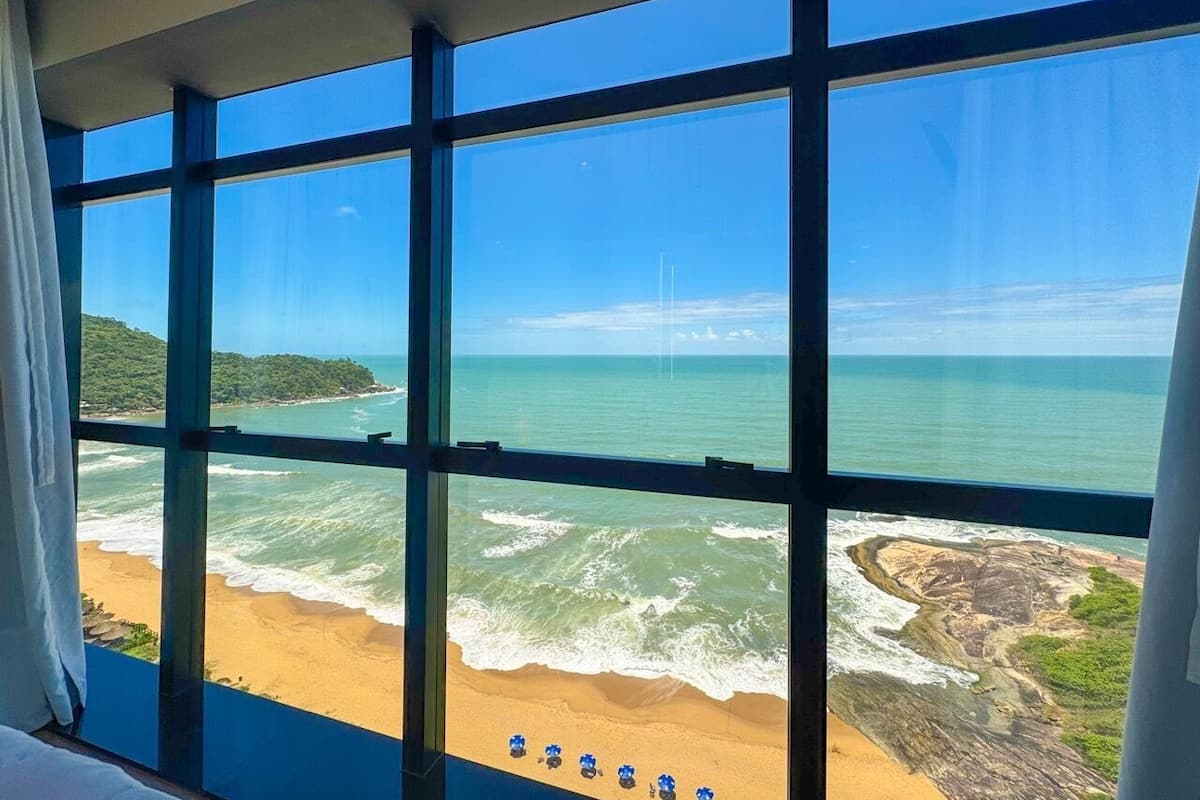 Vista del océano a través de amplios ventanales, con olas rompiendo sobre una playa arenosa y afloramientos rocosos a lo lejos.