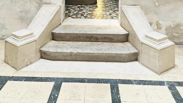 Un escalier avec une rampe en métal mène à une porte. Les murs sont blancs, mais présentent quelques signes d’usure, et le sol est recouvert de carreaux formant un motif.