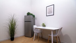 Une petite cuisine équipée d’un réfrigérateur, d’une table blanche et de deux chaises. On trouve une plante en pot à côté du réfrigérateur et un tableau encadré au mur.