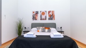 Un lit bien fait, recouvert d’une couverture noire, avec des oreillers blancs et des serviettes pliées. Trois œuvres d’art sont accrochées au mur au-dessus du lit.