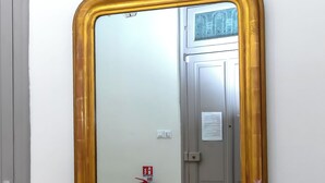 Un miroir avec un cadre doré, accroché au mur, une étagère noire en dessous et un extincteur en arrière-plan.
