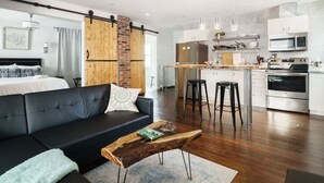 Un salon moderne avec un canapé noir, une table basse en bois et un coin cuisine doté de meubles blancs et d’appareils électroménagers en acier inoxydable.