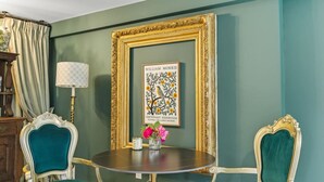 A room with a round table, two chairs, a framed artwork, a lamp, and a vase with flowers.