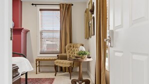 A room with a bed, a bench, a chair, a table with a plant, and a window with blinds.